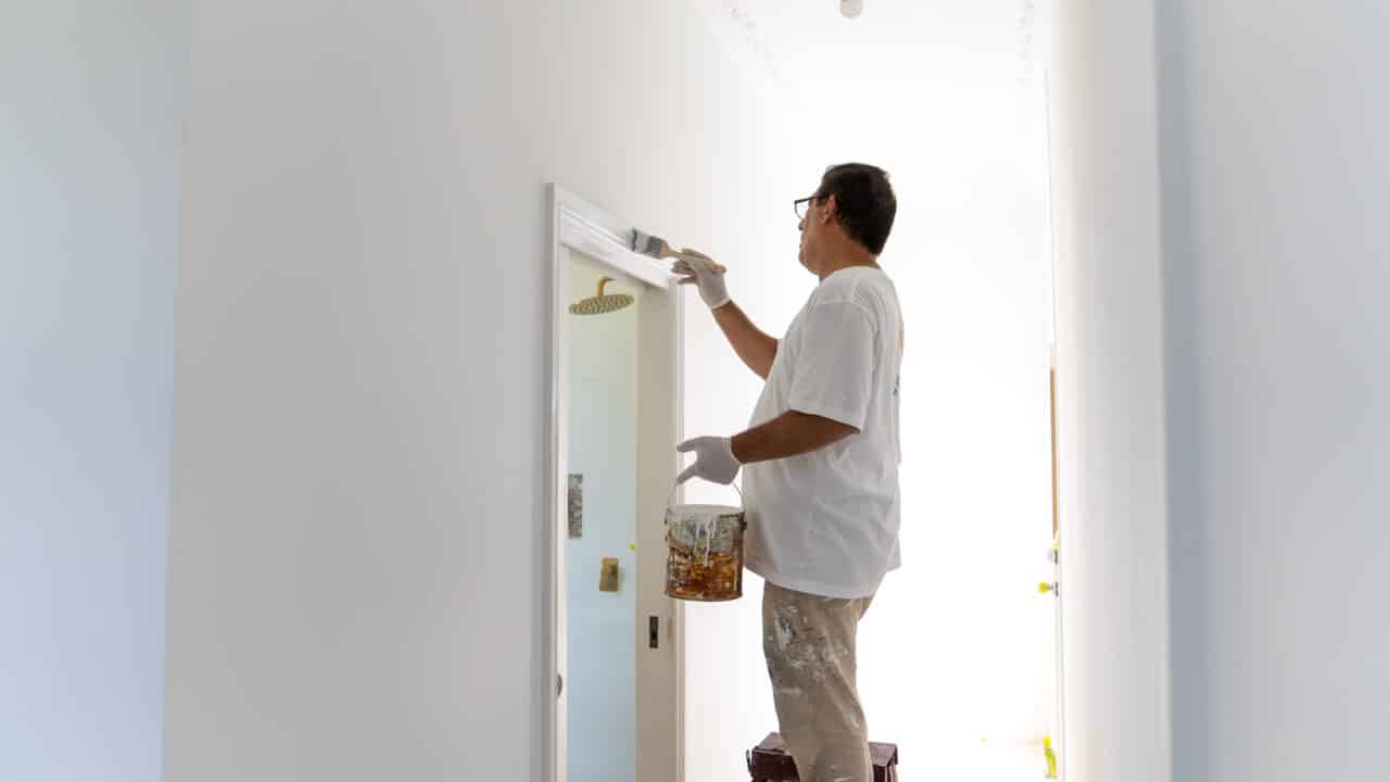 A person wearing a white shirt and gloves stands on a stool, painting a door frame white in a bright hallway, showcasing the quality of Interior Painting Services available in the Blue Mountains.