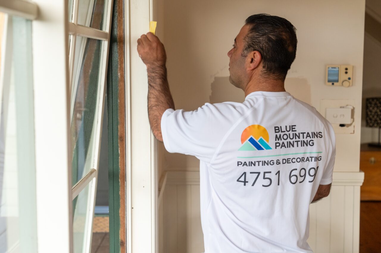 A person wearing a Blue Mountains Painting shirt is preparing to paint a wall, holding a scraper tool near a doorway, showcasing the meticulous approach of professional painters.