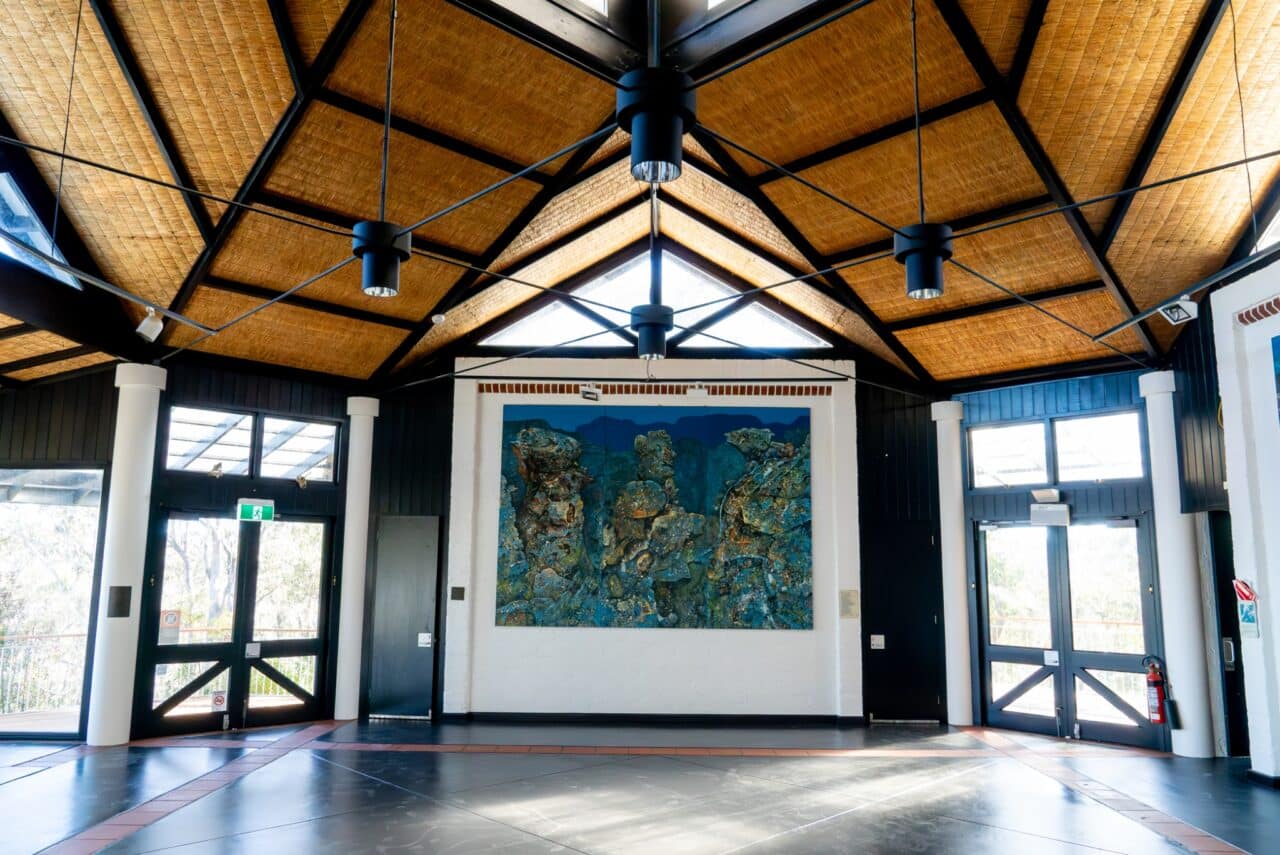 An interior shot of a spacious hall with a high ceiling, featuring a large abstract painting on the far wall, ceiling fans, and natural light streaming through windows near the entrance and side walls evokes a sense of conservation with its eco-friendly design while also maintaining the cozy charm of a modern hut.