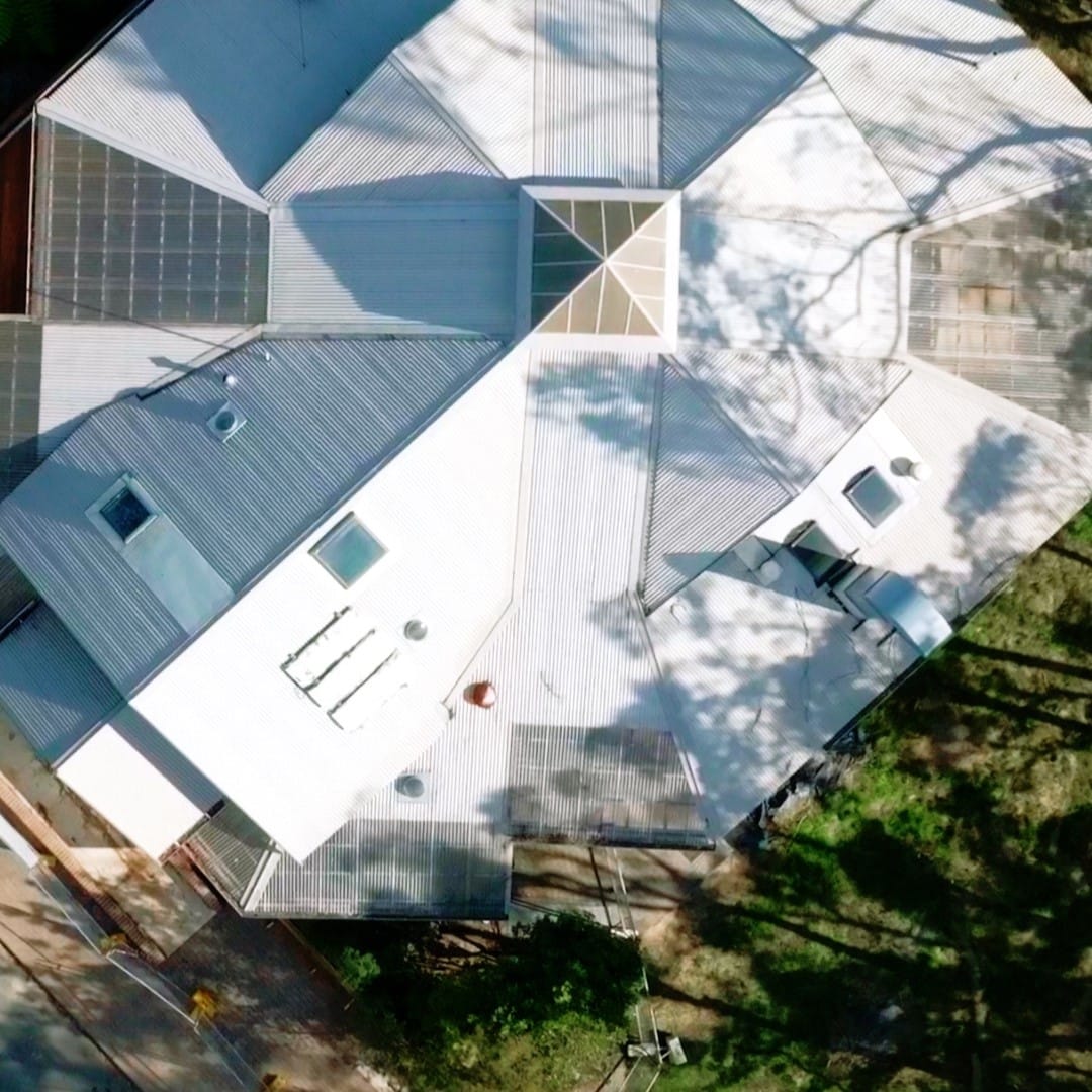 Aerial view of a unique, multi-faceted building with a metallic roof and solar panels on several sections, blending modern architecture with conservation efforts, surrounded by trees and shadows.