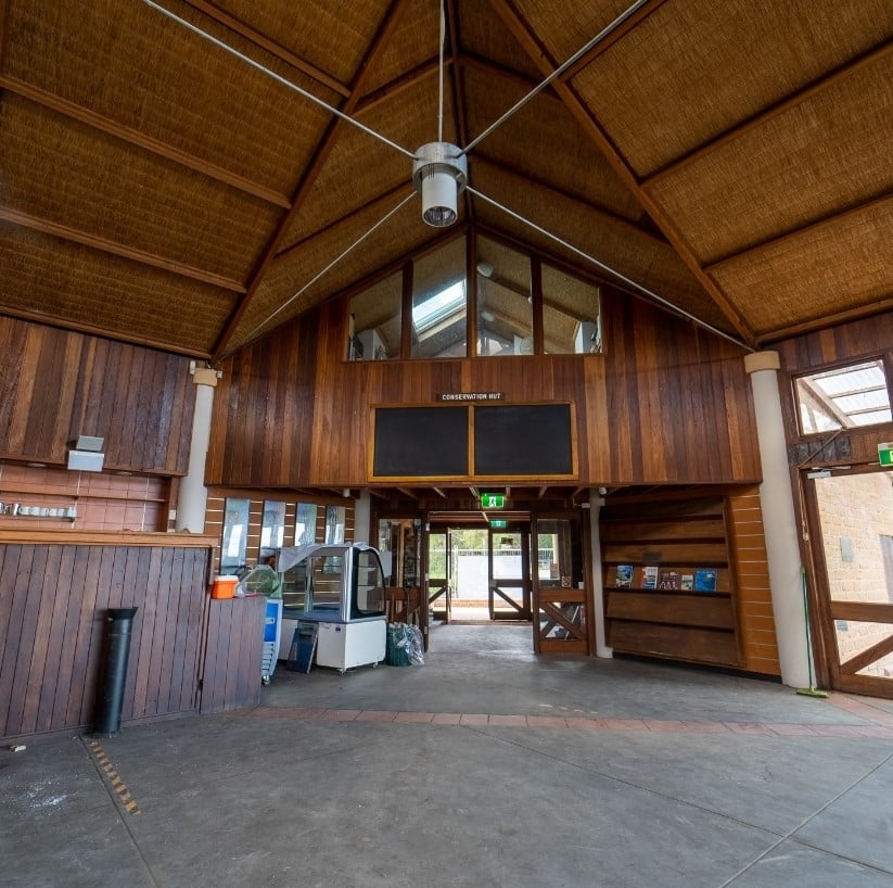 A spacious wooden interior with high ceilings, reminiscent of a rustic hut, featuring large windows, a vending machine, and an exit door with a green emergency sign above it.