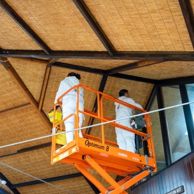 Two workers in white uniforms stand on an orange scissor lift, meticulously conserving the wooden ceiling of a hut-like building.