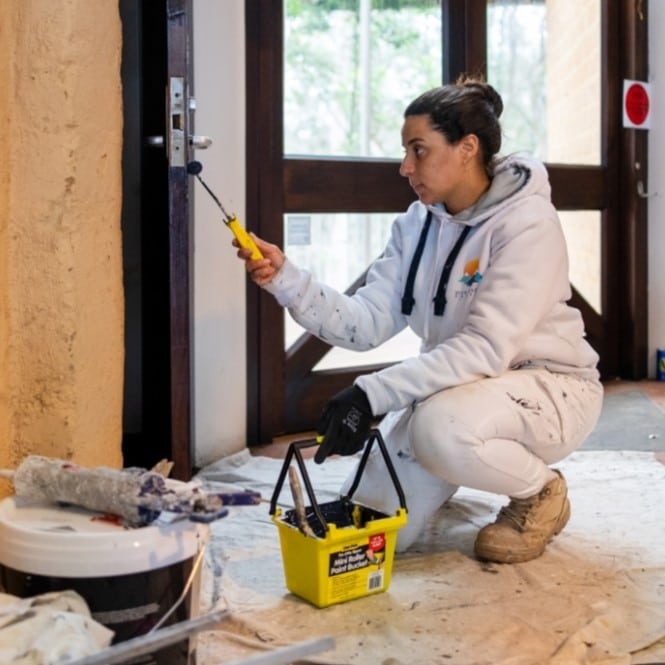 A person in white work attire kneeling while painting a door with a small roller, holding a yellow paint bucket with black handles, surrounded by painting supplies on a drop cloth, appears focused as though working on the conservation of an old hut.