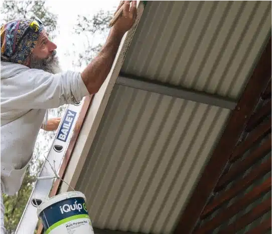 A person with a colorful headscarf, reminiscent of the vibrant hues of the Blue Mountains, paints the underside of a corrugated roof while standing on a ladder, with a paint bucket hanging nearby.