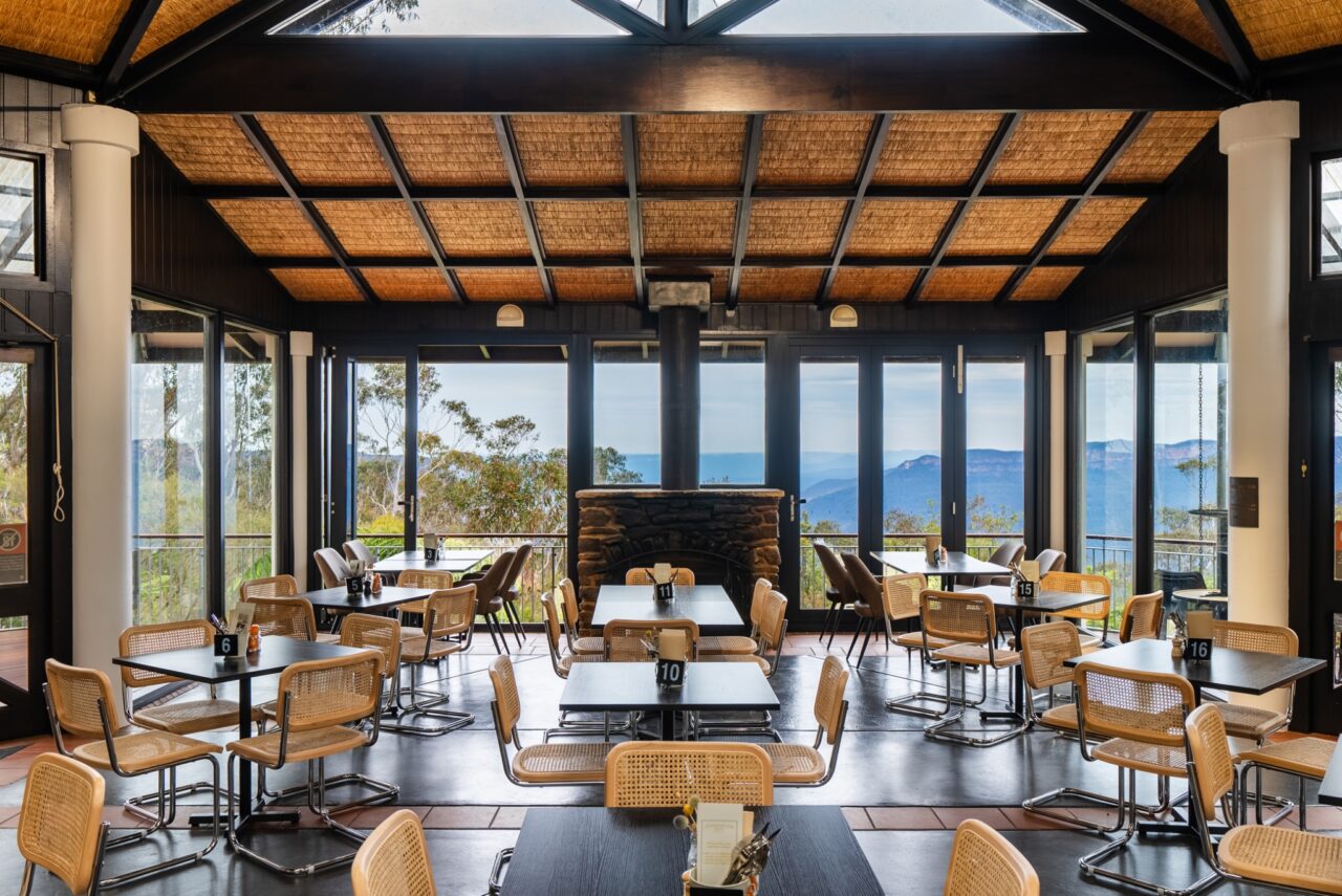 A spacious dining area with large windows, a wooden ceiling, wicker chairs, set tables, and a panoramic view of mountains and trees evokes the charm of a conservation hut.