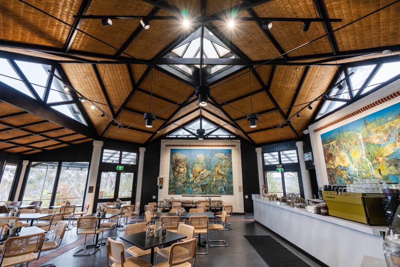 A modern café with a wooden ceiling reminiscent of a cozy hut, featuring skylights, multiple tables and chairs, and a counter on the right. A large mural decorates the far wall.