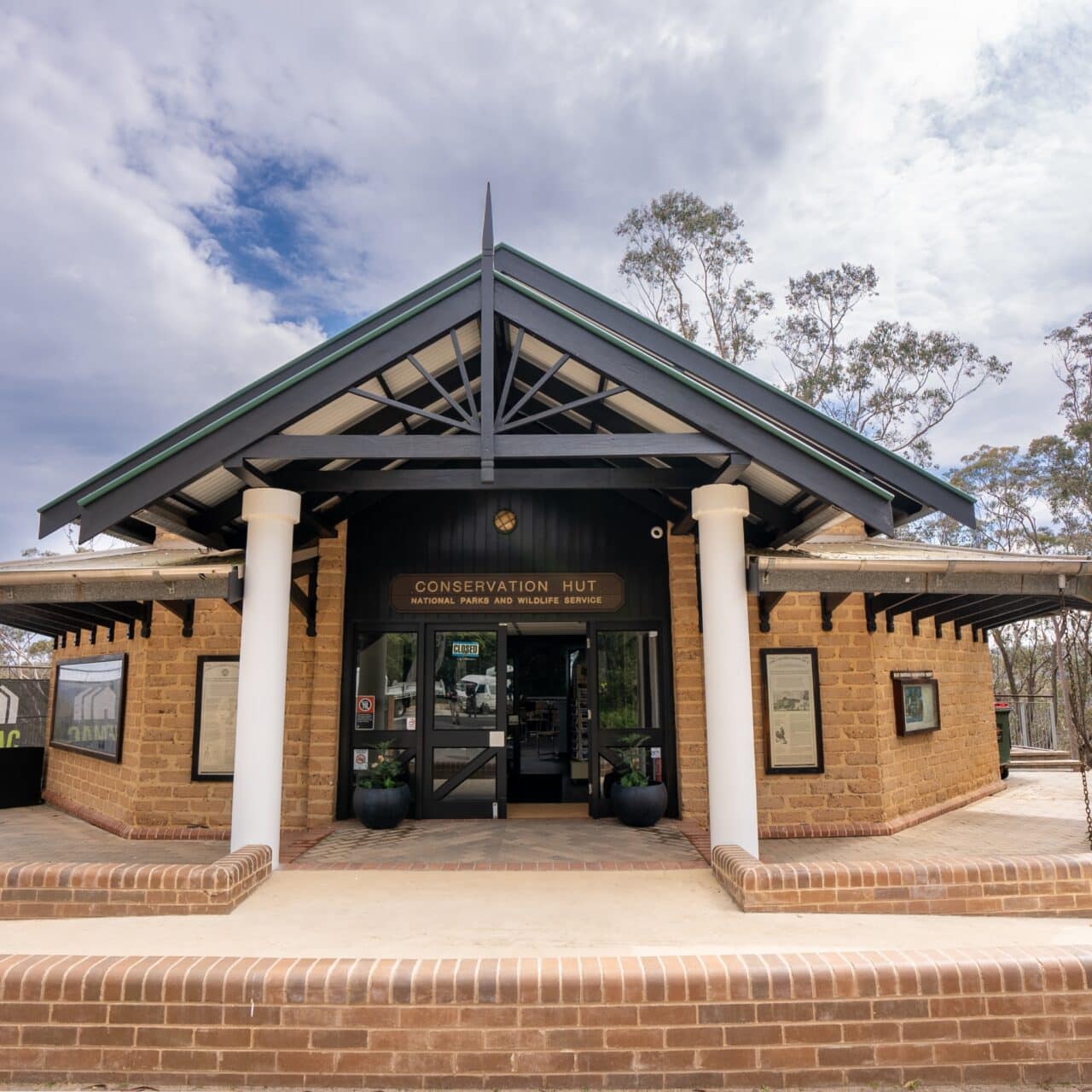 A brick building with an arched entrance labeled "Conservation Hut" under a metal roof, framed by trees and a partly cloudy sky.