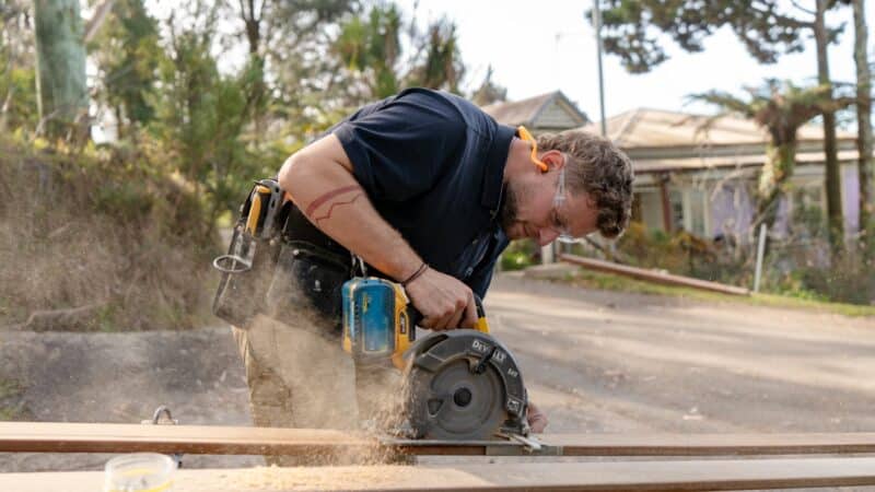 Person using a circular saw to cut wood outdoors, wearing safety glasses and a tool belt, with trees and a house in the background.