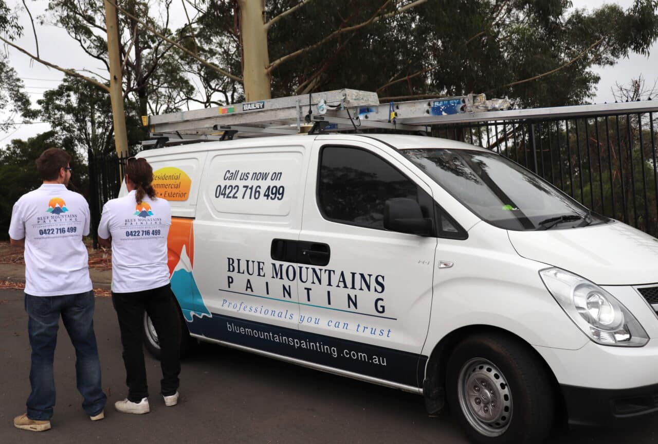 Two people in branded shirts standing next to a Blue Mountains Painting van with ladders on the roof.