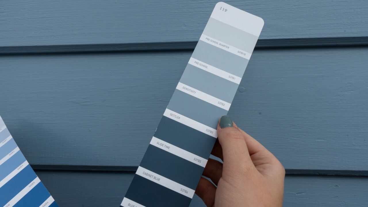 Hand holding a blue paint swatch card with shades labelled Pre School Quarter, Sensitivity, Settler, Glide Time, and Cabinet Blue, against a matching blue-painted wall.