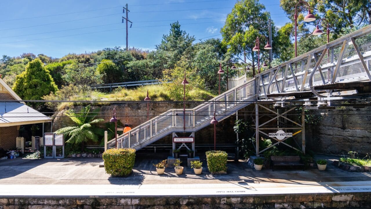 A small train station platform with metal stairs leading to an upper level, signs display "Mount Victoria." Several potted plants and trees are visible, with greenery in the background. The recent restoration has enhanced the charm of Mt Vic Train Station.