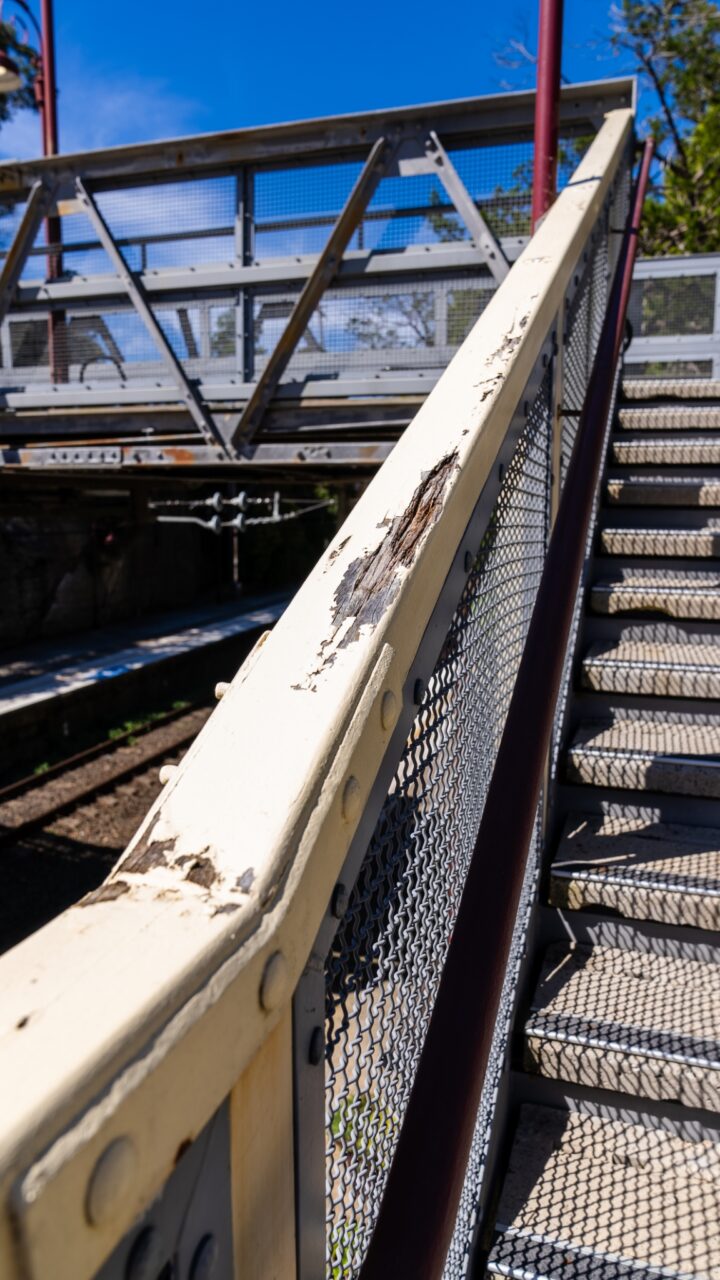 A close-up of a metal staircase with peeling paint on the handrail, leading up to a pedestrian bridge at Mt Vic Train Station. The sky is clear and blue in the background, hinting at potential restoration projects ahead.