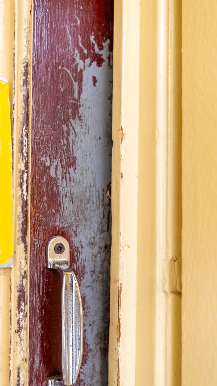 Close-up of an old, chipped door frame painted yellow with a metal handle. Adjacent is a wooden panel showing signs of wear and paint peeling, reminiscent of the charm at Mt Vic Train Station in need of restoration.