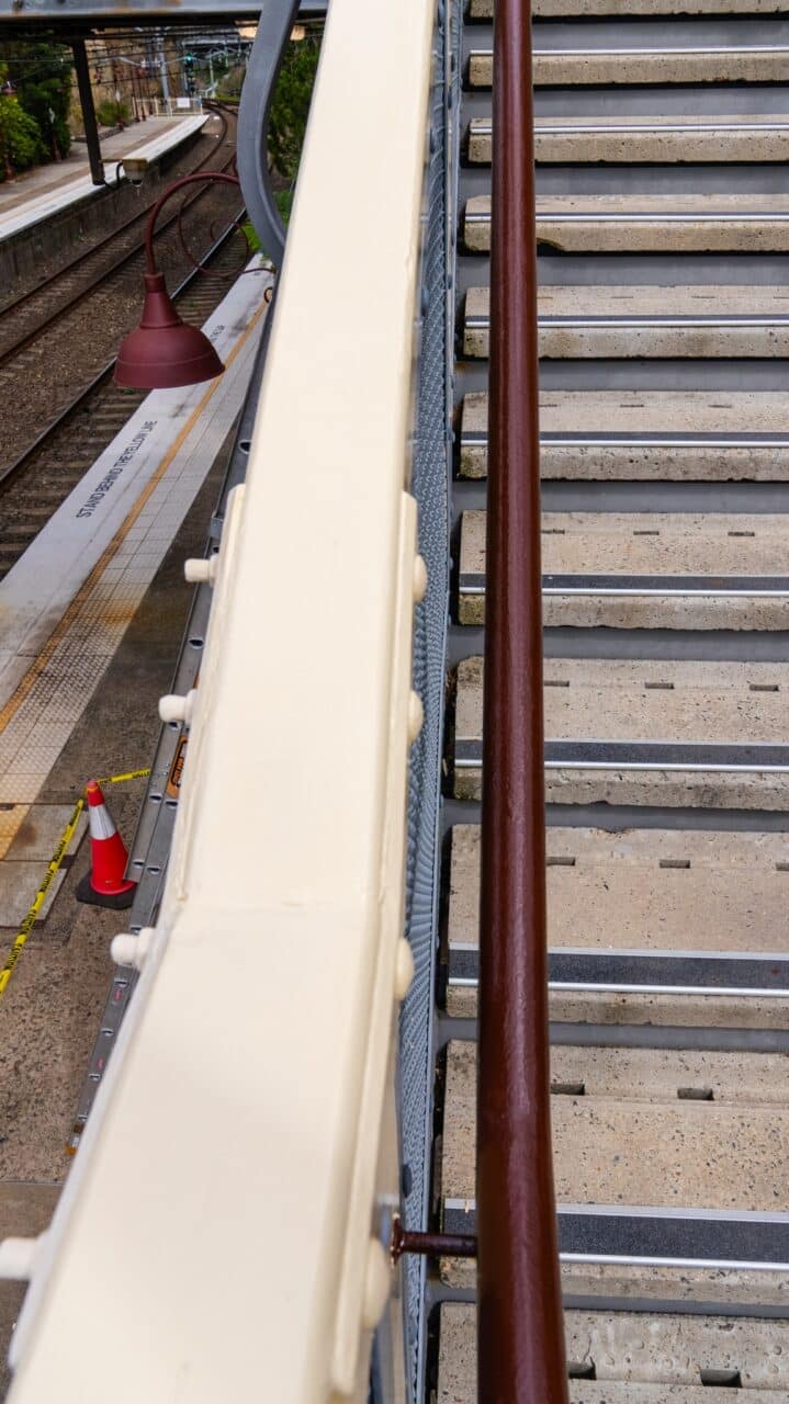 A staircase beside the Mt Vic Train Station platform features beige handrails, with a brown handrail adjacent to them. Warning cones are visible on the platform as part of the ongoing restoration efforts.