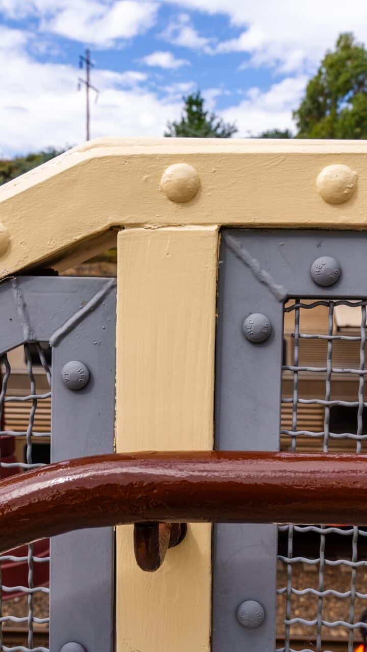 Close-up of a painted metallic structure with cream-colored beams, grey mesh, and a rust-colored handrail at Mt Vic Train Station. Trees and sky with clouds visible in the background tell of its meticulous restoration.