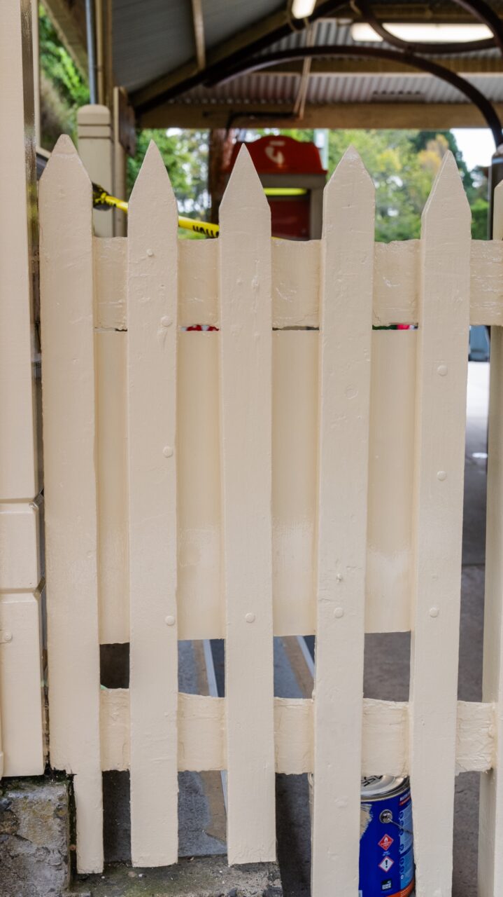 A white wooden picket fence stands with a paint can and brush behind it, indicating ongoing restoration work. Yellow caution tape adds a cautionary note, reminiscent of the meticulous care often seen at places like Mt Vic Train Station.