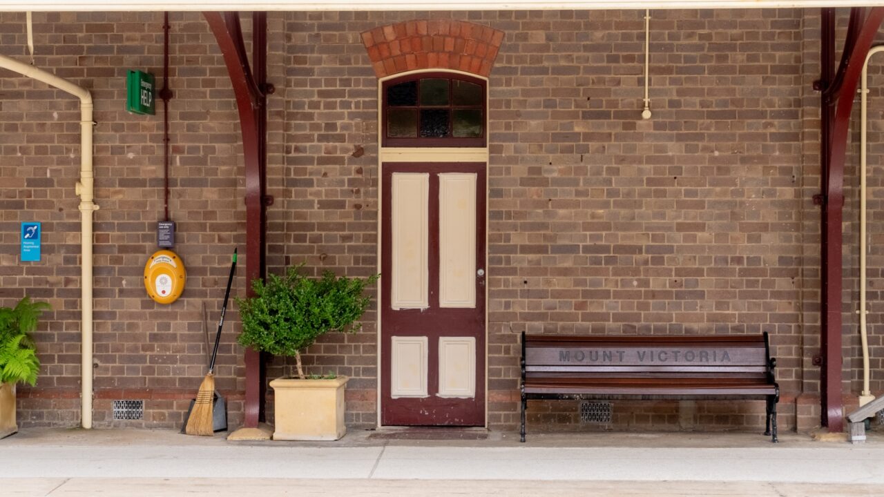 A brick wall with a white door, a bench labeled "Mount Victoria," a potted plant, broom, and emergency equipment evoke the charm of the Mt Vic Train Station under restoration.