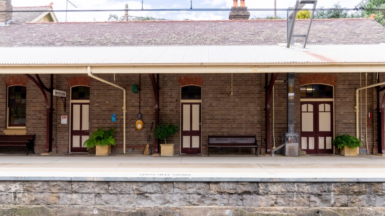 Empty train platform with four red-and-white doors, brick building, plants, a bench, and an overhead shelter. The station is named "Mt Vic Train Station," with a visible sign highlighting its recent restoration.