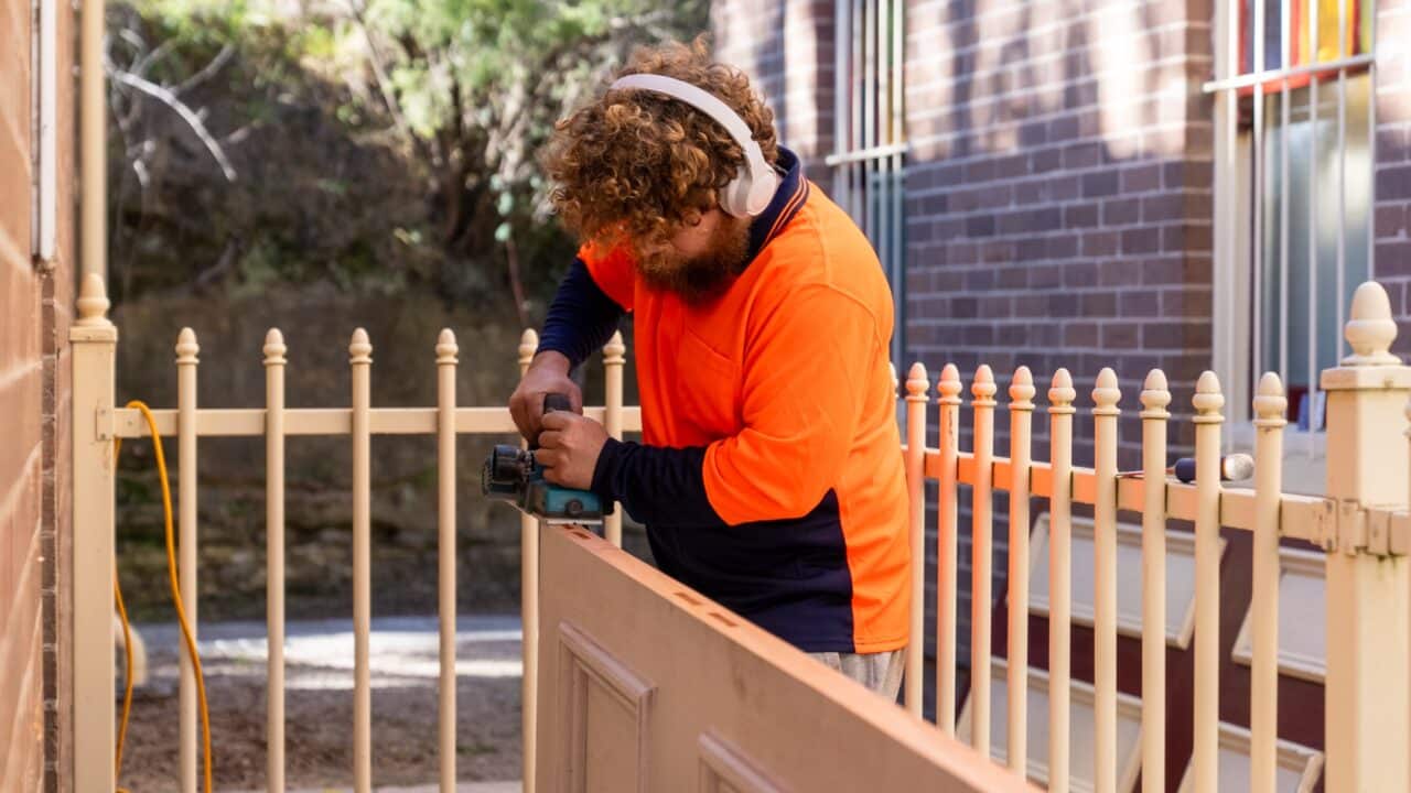 A person wearing an orange and navy shirt uses a power tool on a wooden board outside, next to a beige metal fence near the Mt Vic train station. The person, working on what appears to be a restoration project, is wearing headphones and has curly hair.