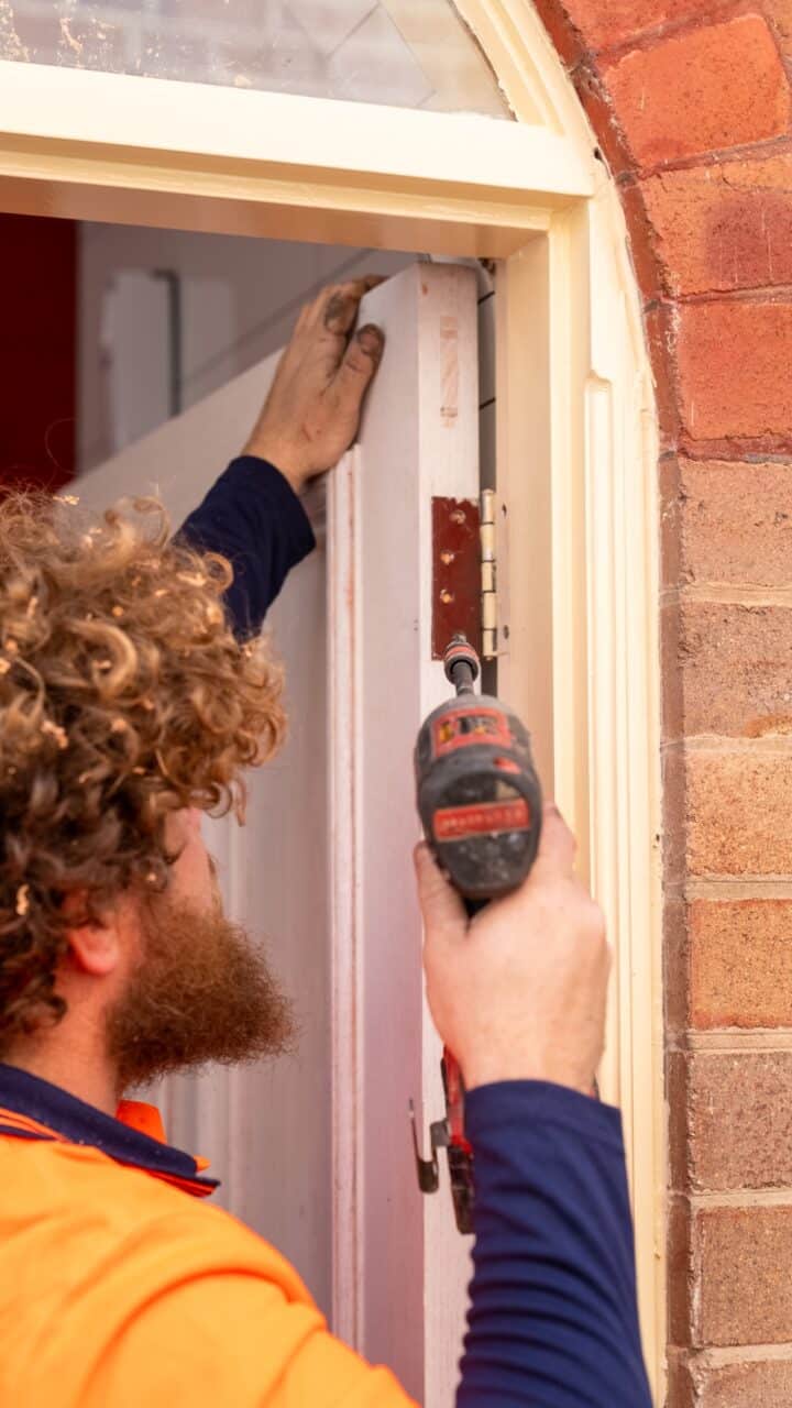 A worker with curly hair installs a door hinge using a power drill near a brick wall during the restoration of Mt Vic Train Station.