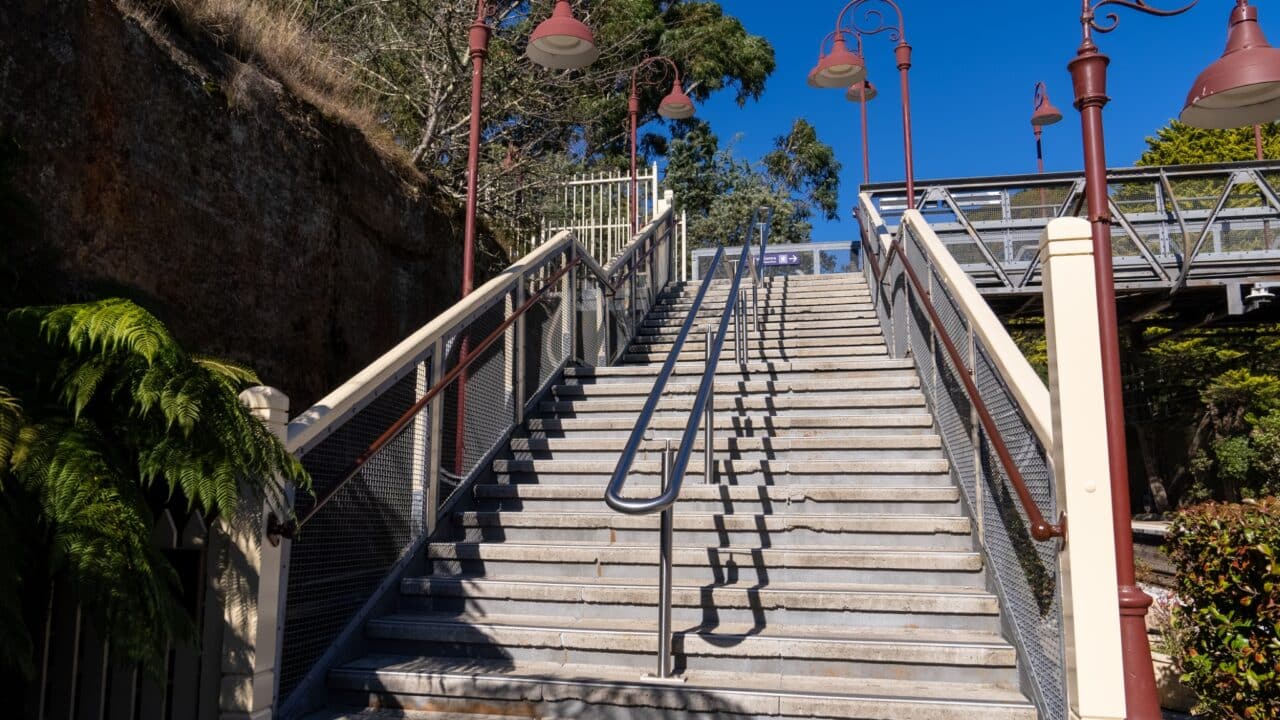 A stone staircase with metal railings, part of the recent restoration, leads up towards a platform under a clear blue sky at Mt Vic Train Station.