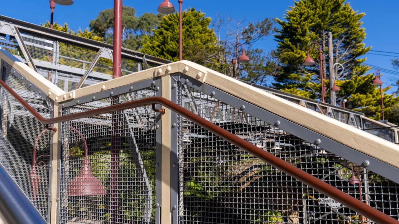 View of an outdoor staircase with metal railings and a wooden handrail, surrounded by trees and under a clear blue sky, reminiscent of the thoughtfully designed spaces at the restored Mt Vic Train Station.