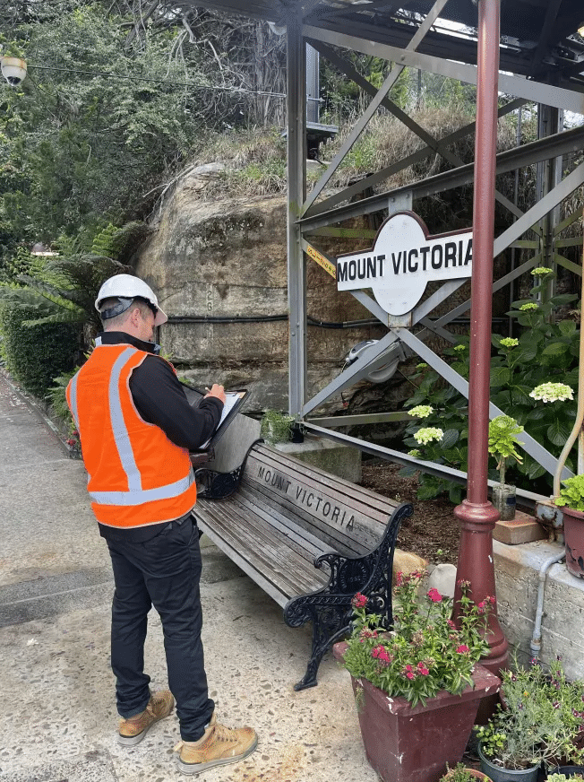 A person wearing an orange safety vest and white helmet writes on a clipboard near a bench and sign labeled "Mt Vic" beside a rocky wall and greenery, likely part of the ongoing restoration project.