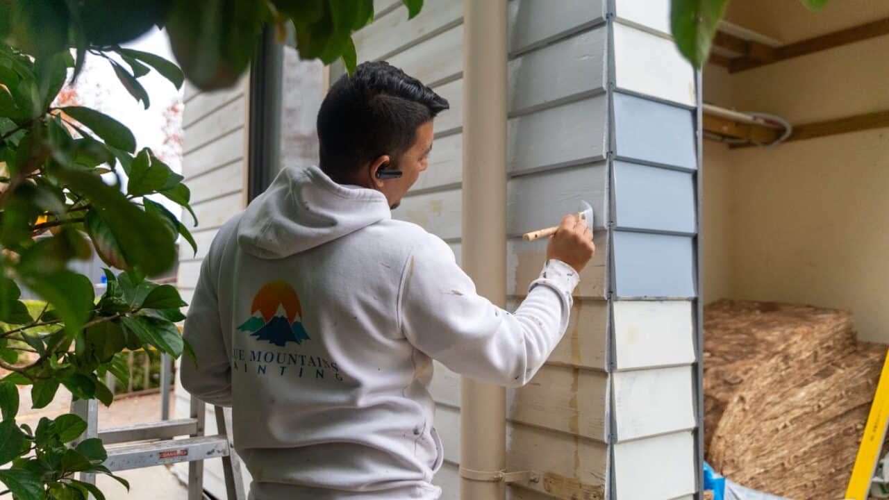 A person wearing a hoodie with a painting company logo is providing exterior painting services, carefully painting a house exterior blue using a paintbrush.