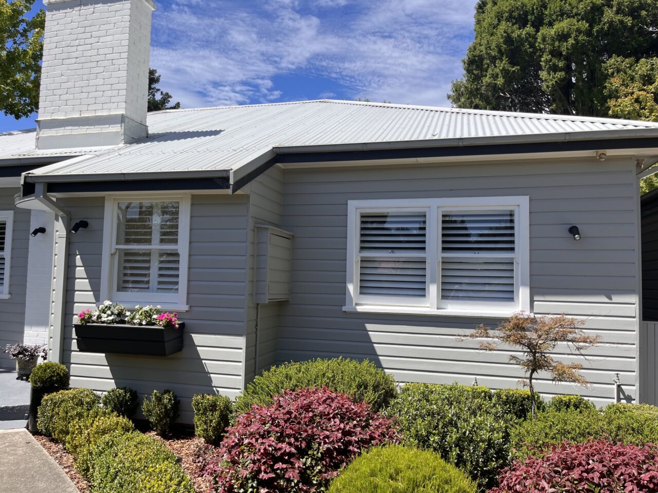 A small, single-story house with light gray siding, white trim, and a metal roof, surrounded by manicured shrubs and a flower bed. White shutters cover the windows. Strata Painting Services recently refreshed the exterior, enhancing its charm amidst the stunning Blue Mountains backdrop.