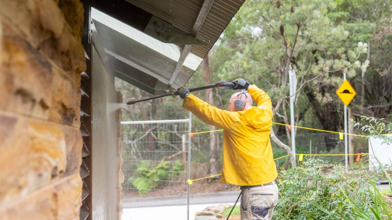 A person in a yellow jacket power washes the side of a building. They are outdoors with greenery and a road sign visible in the background, showcasing Professional Exterior House Cleaning services near the Blue Mountains.