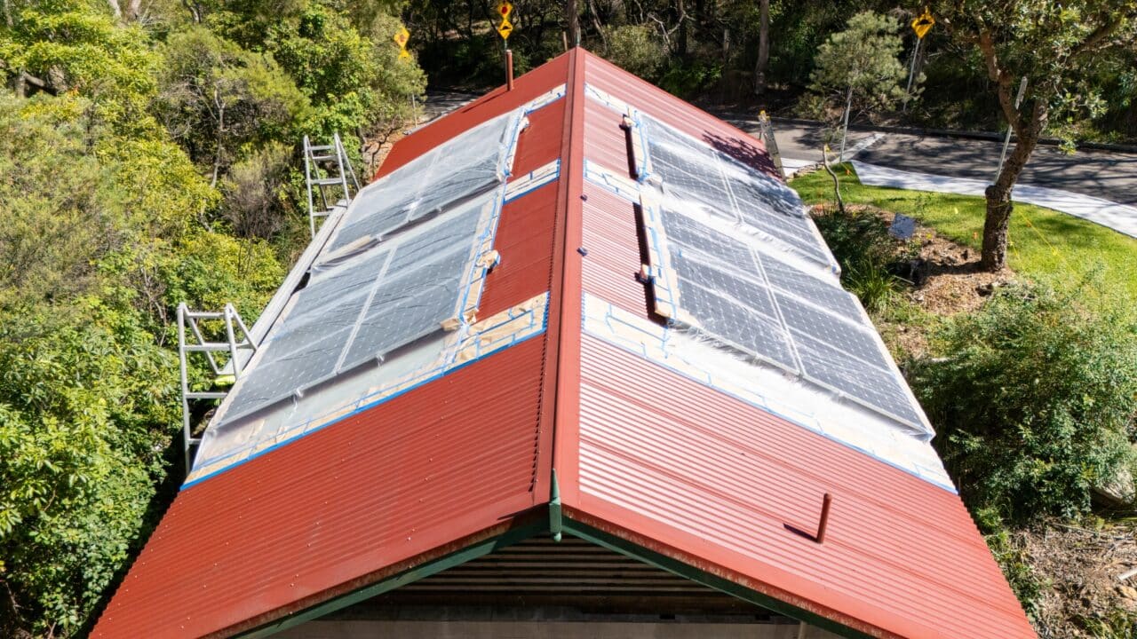 A red-roofed building with partially installed solar panels, surrounded by trees and a road in the background. Two ladders are placed against the roof, indicative of ongoing Roof Restorations Blue Mountains projects.