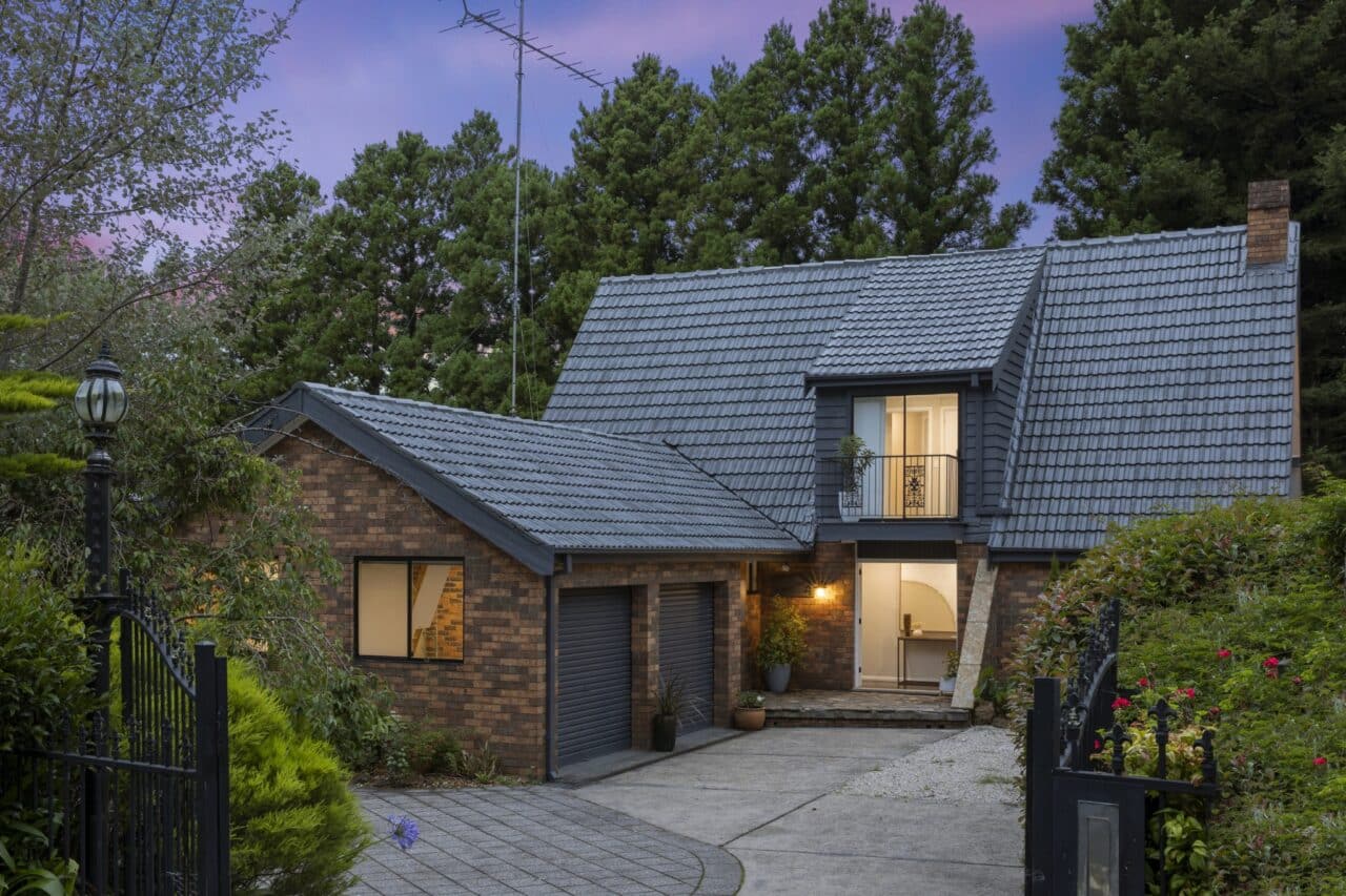 A two-story brick house with a gray tiled roof, surrounded by trees in the Blue Mountains. The house features a two-car garage, front yard, and a lit porch area. The evening sky is visible in the background, highlighting the recently finished work of quality roof painting services.