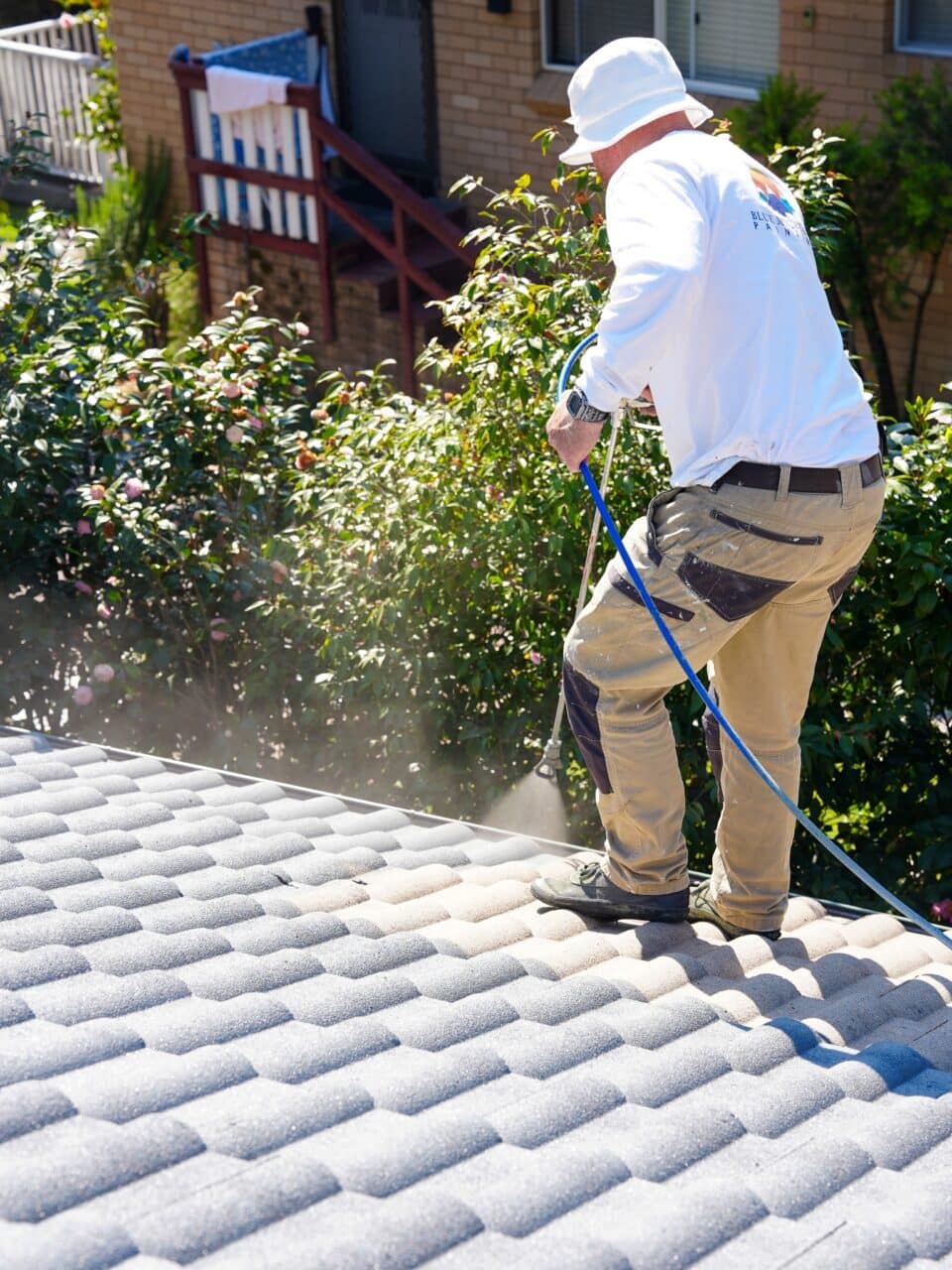 A person in work clothes and a hat is using a hose to clean a tiled roof near a garden with bushes, exemplifying professional exterior house cleaning in the Blue Mountains.