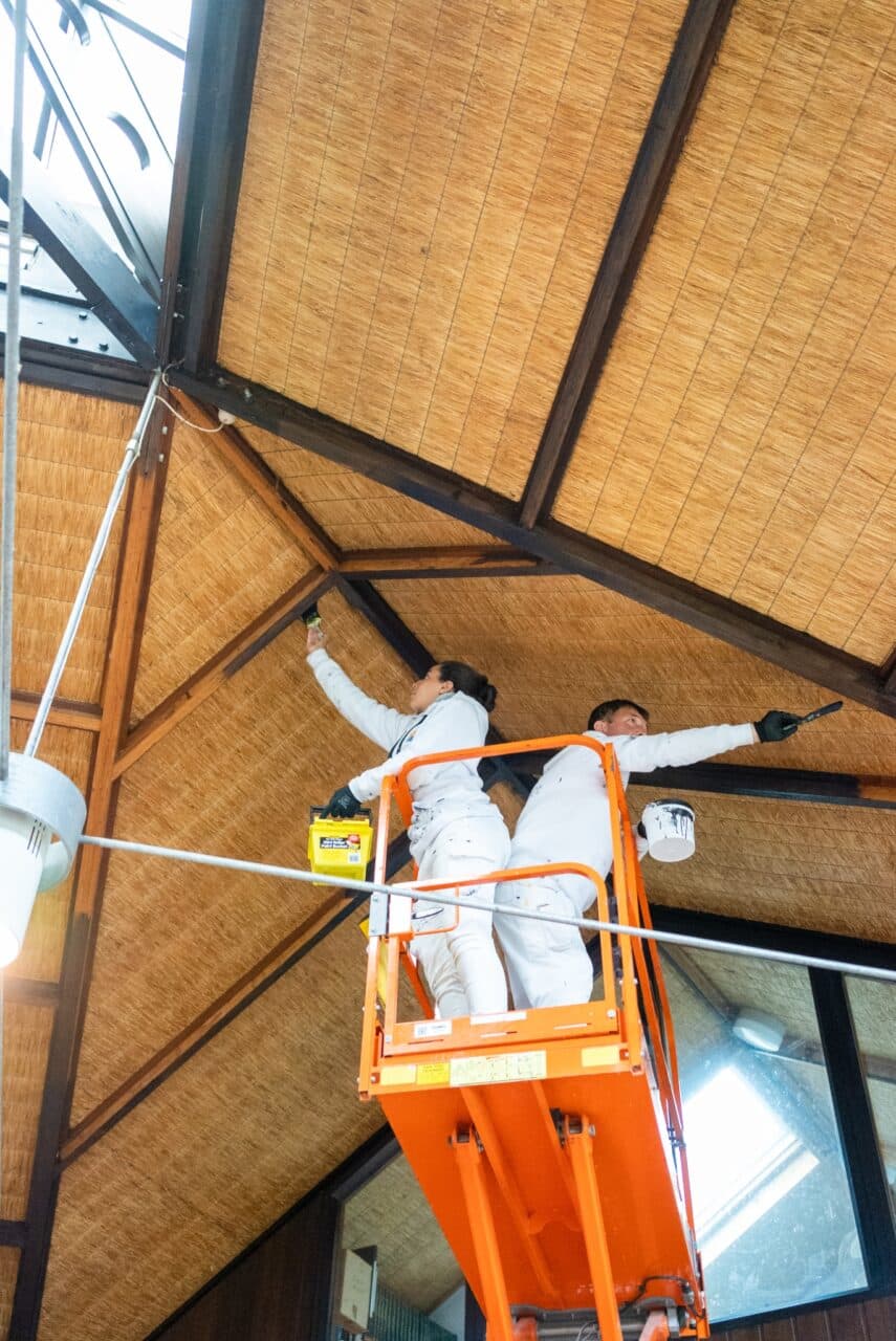 Two individuals in white overalls on an orange scissor lift are painting the wooden ceiling of a high room. One holds a paint can while the other applies paint with a brush, showcasing their expertise as part of Commercial Painting Services in the Blue Mountains.