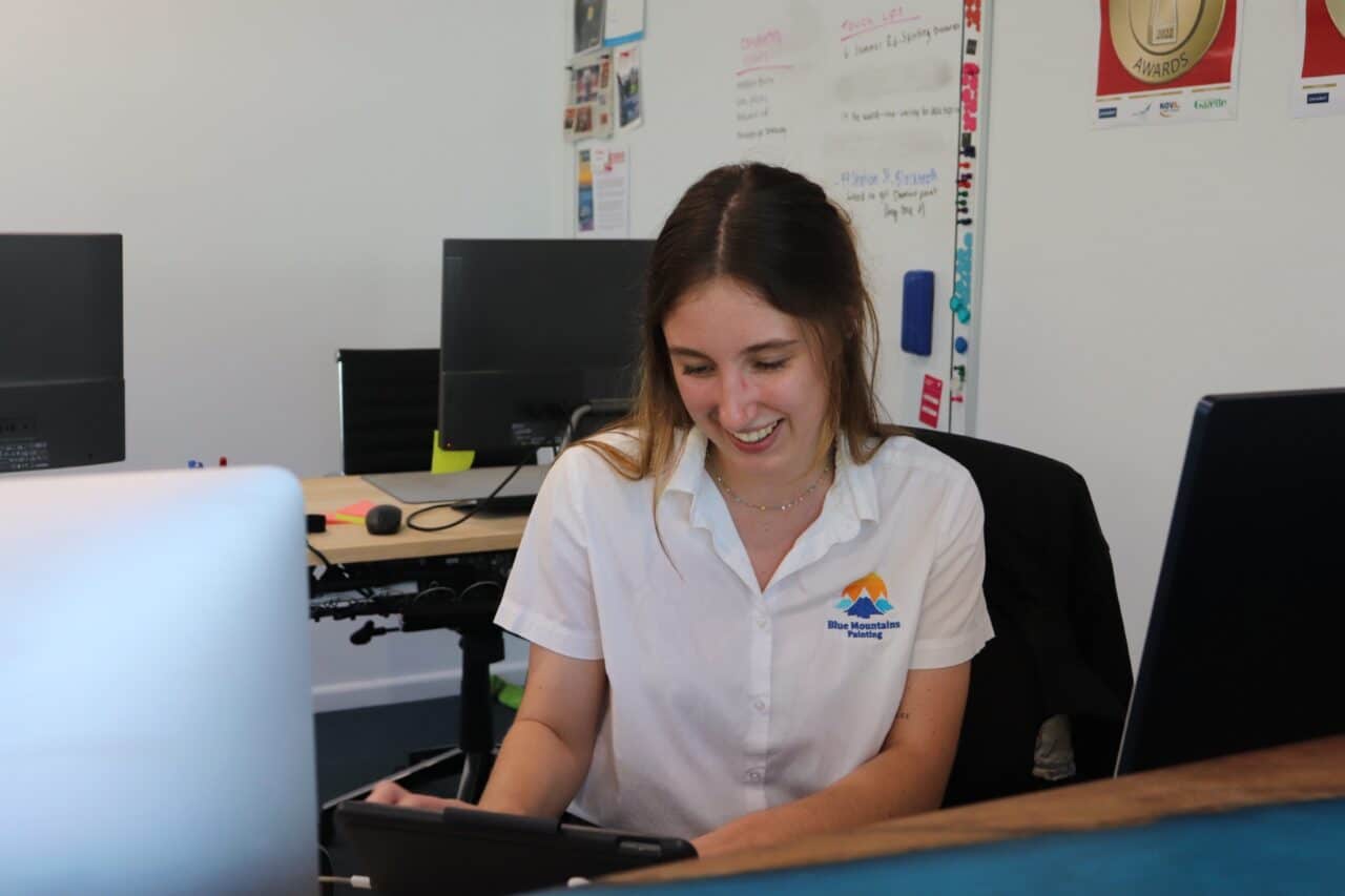 A woman in a white blouse sits at a desk, smiling and using a tablet. Office environment with laptops and whiteboard in the background.