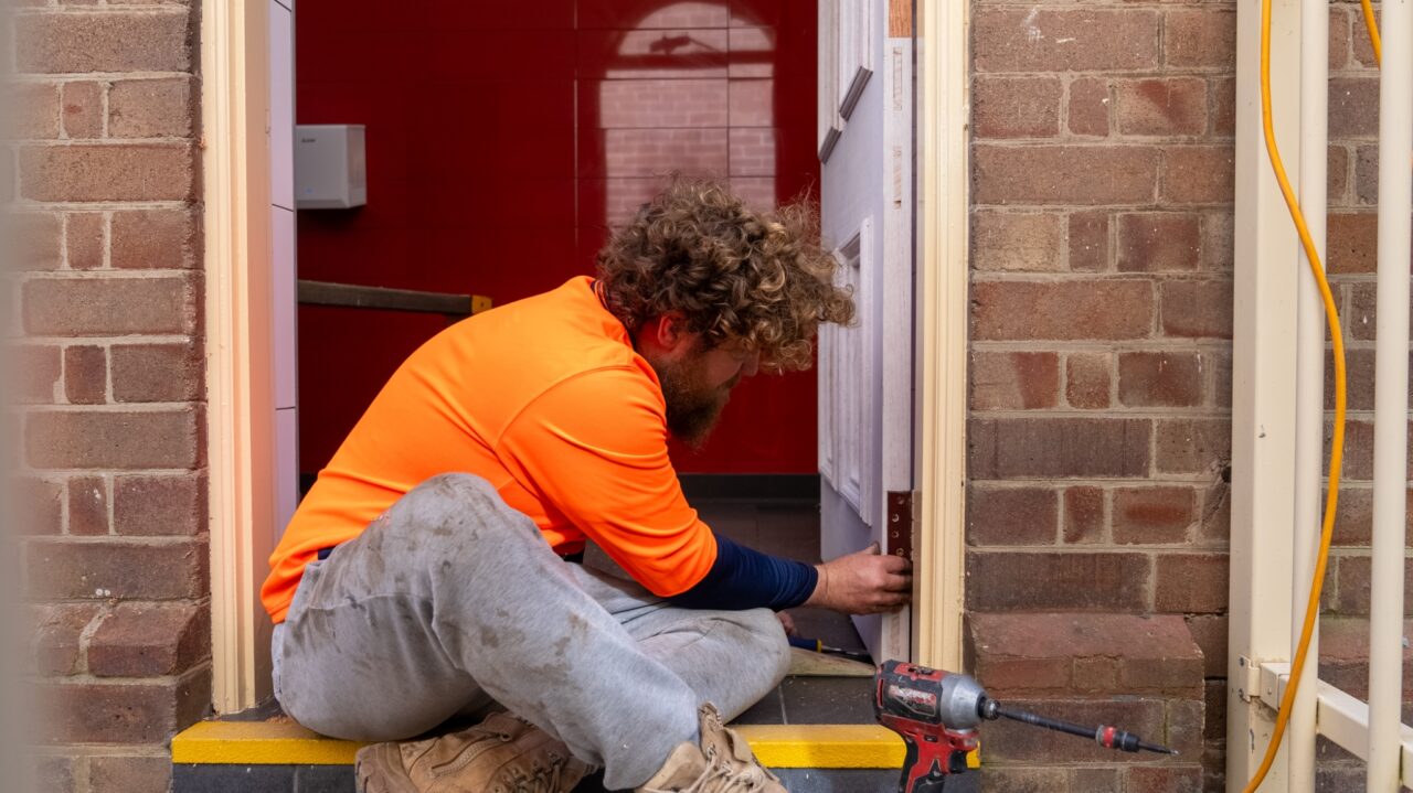 A man wearing an orange shirt and gray pants is seated while installing a door frame in a brick-walled structure. A drill is placed beside him, showcasing the precision of Handyman Services.