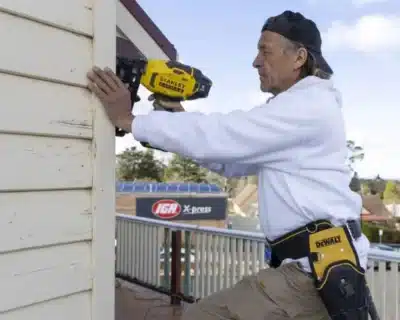 A man in a white hoodie and backwards cap uses a nail gun on an exterior wall. Sporting a tool belt, he works diligently with the precision you’d expect from Professional Painters. In the background, an "IGA X-press" store sign stands out against the picturesque Blue Mountains.