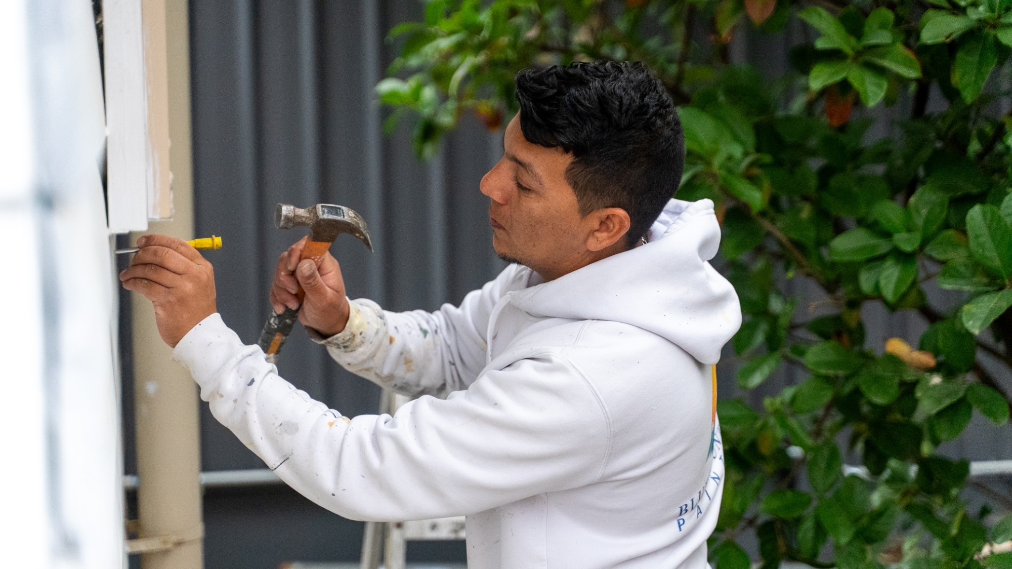 A person in a white hoodie uses a hammer and a yellow measuring tool to work on an outdoor structure with greenery in the background, reminiscent of the professional painters often seen around the scenic Blue Mountains.