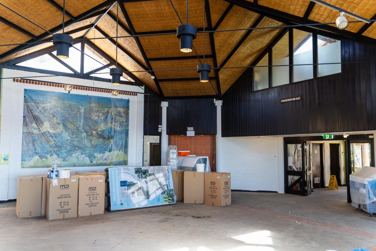 Interior of a hut with high ceilings exposing structural beams, stacked cardboard boxes, and a large mural on the wall. The room appears unfinished or in the process of being set up, reflecting an emphasis on conservation through simple yet functional design.