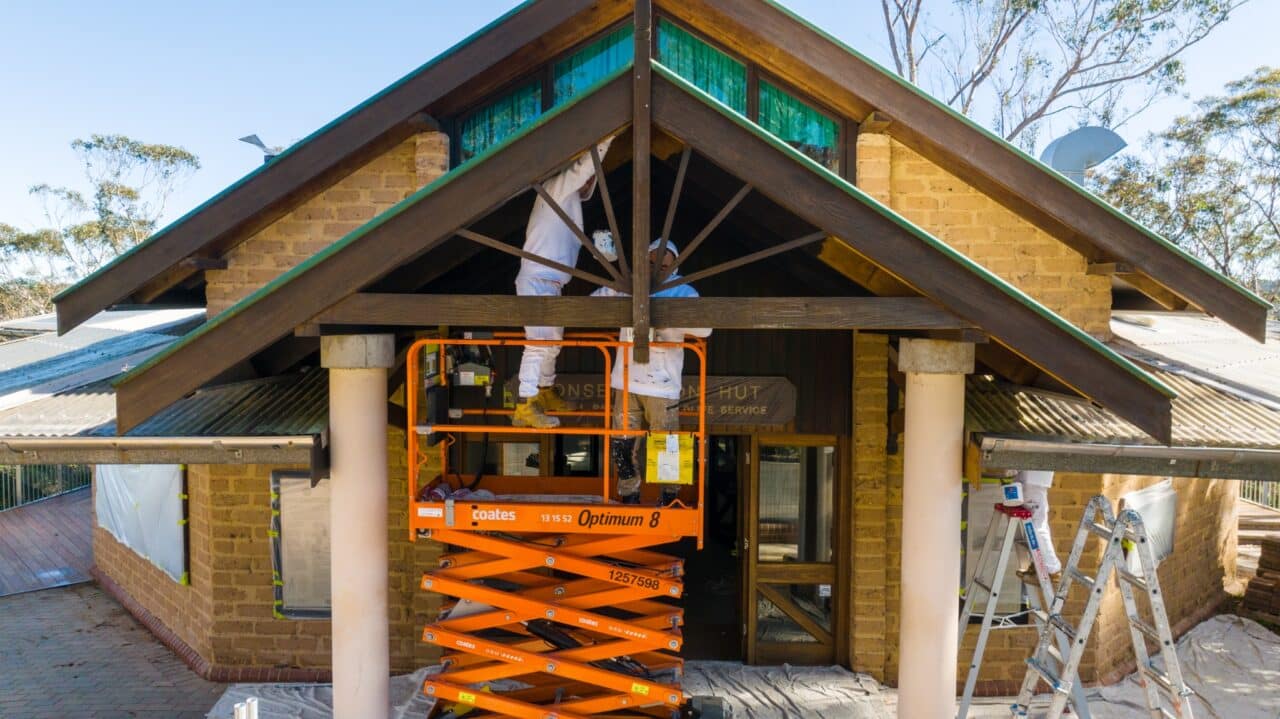 Two workers in white protective suits are using a scissor lift to conserve the exterior of a house with a brown brick facade. Positioned between two roof gables, they work diligently, with ladders on each side, ensuring every detail is meticulously attended to.