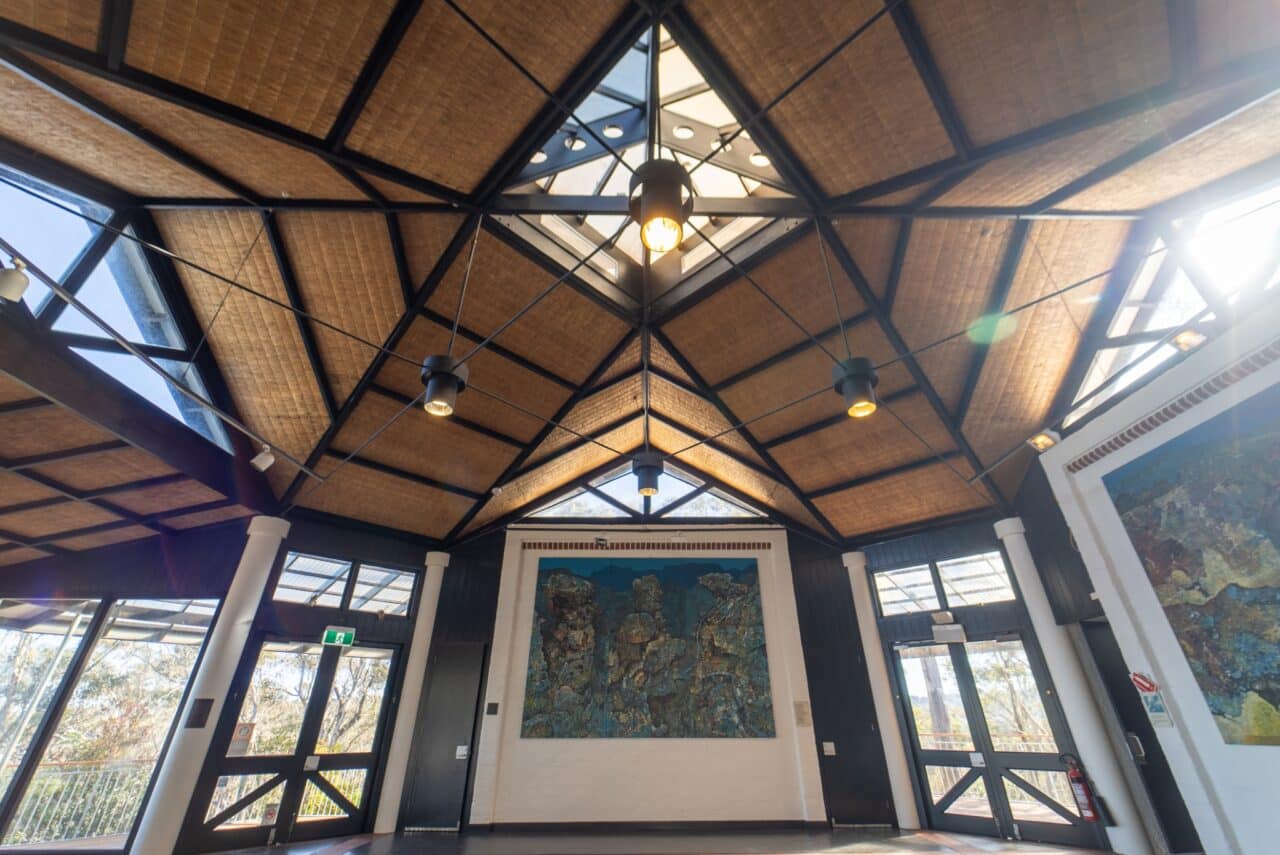 A spacious room with a high, triangular wooden ceiling reminiscent of a hut, large windows, and prominent artwork on a wall. Natural light fills the area, creating a sense of conservation and tranquility.