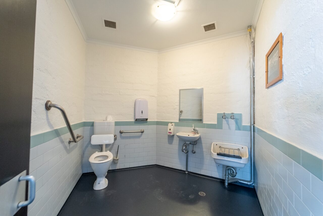 A clean, spacious public restroom with a toilet, sink, soap dispenser, wall-mounted mirror, hand dryer, grab bars, and tiled walls embodies the spirit of conservation often seen in eco-friendly huts.