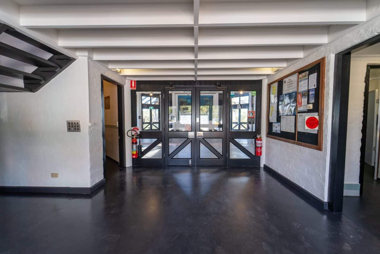 A hallway with a dark floor, white walls, a staircase on the left, and a glass double door exit in the center. A fire extinguisher and bulletin board supporting conservation efforts are on the right wall.