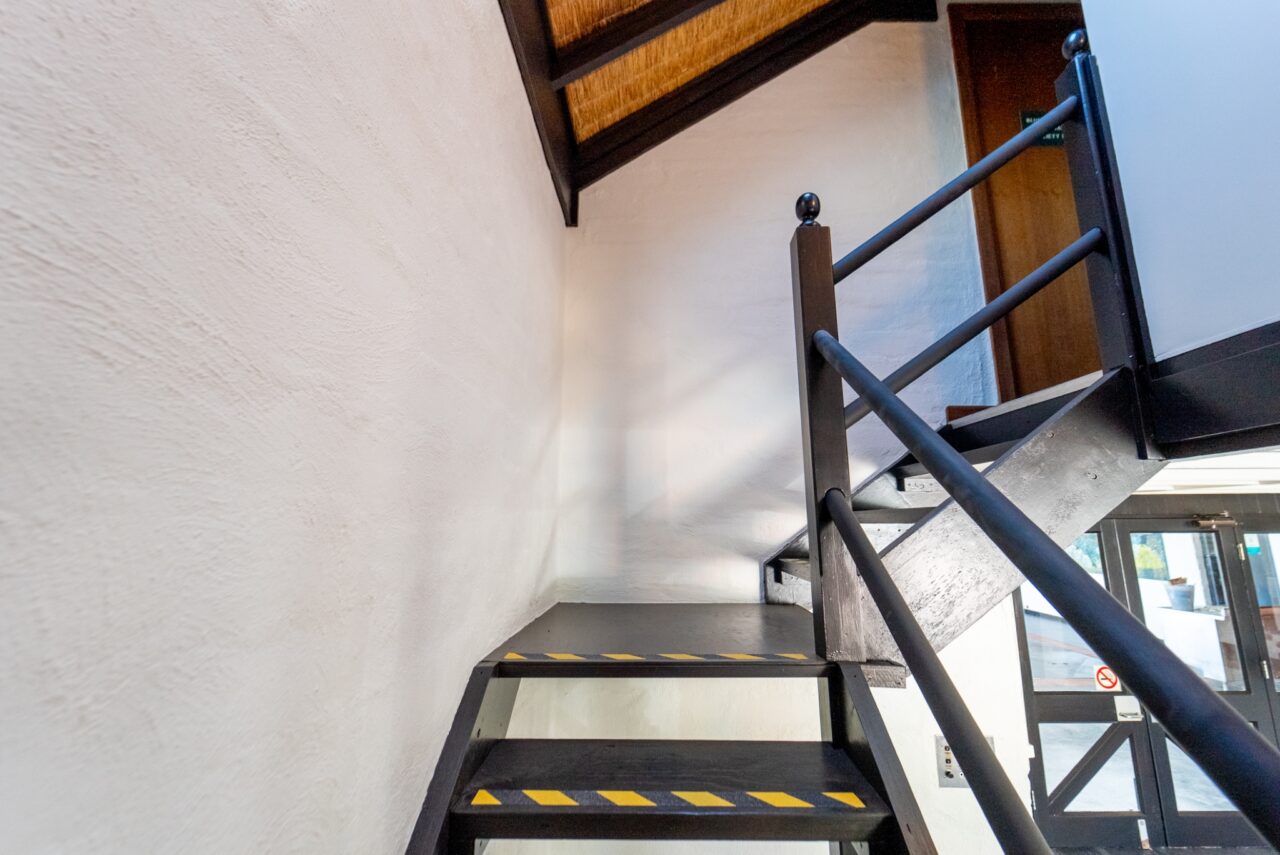 Black metal staircase with yellow safety stripes leads to a loft. White textured wall on the left and a glass door is visible at the top right corner, reminiscent of a conservation hut's simple yet practical design.