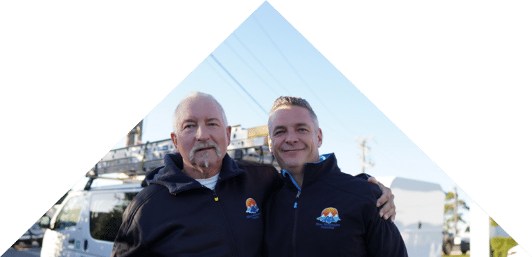 Two men standing side by side, wearing matching jackets with a logo. Behind them, there's a white van with a roof rack parked near a quaint hut. The sky is clear and blue, hinting at their next conservation adventure.