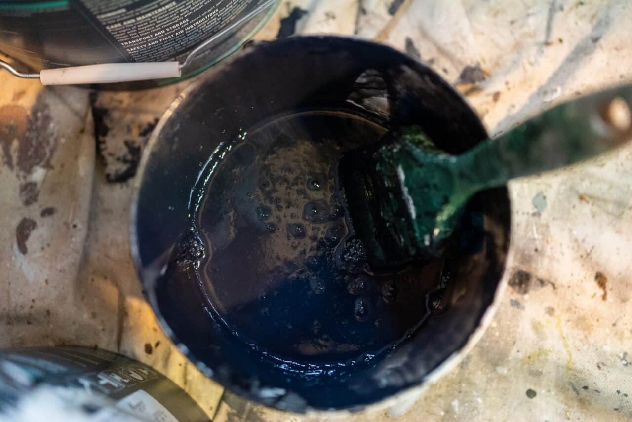 A paintbrush resting inside a container filled with dark blue paint. The container and surrounding area of this artist's hut are covered with paint splatters.