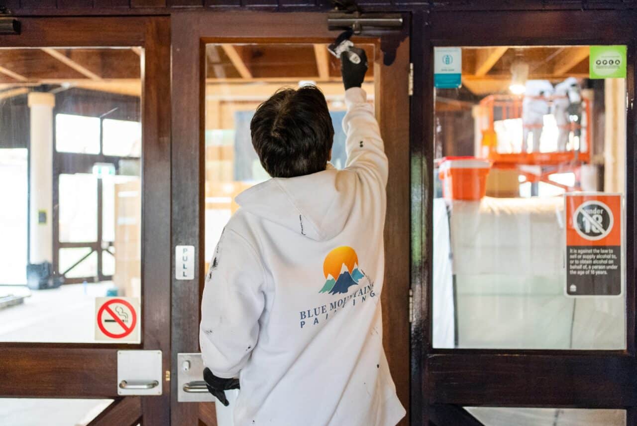 A person in a "Blue Mountain Painting" hoodie paints a wooden door indoors, contributing to conservation efforts. Various signs, including a no smoking sign, are visible on the glass panes of the door. Construction equipment is in the background.