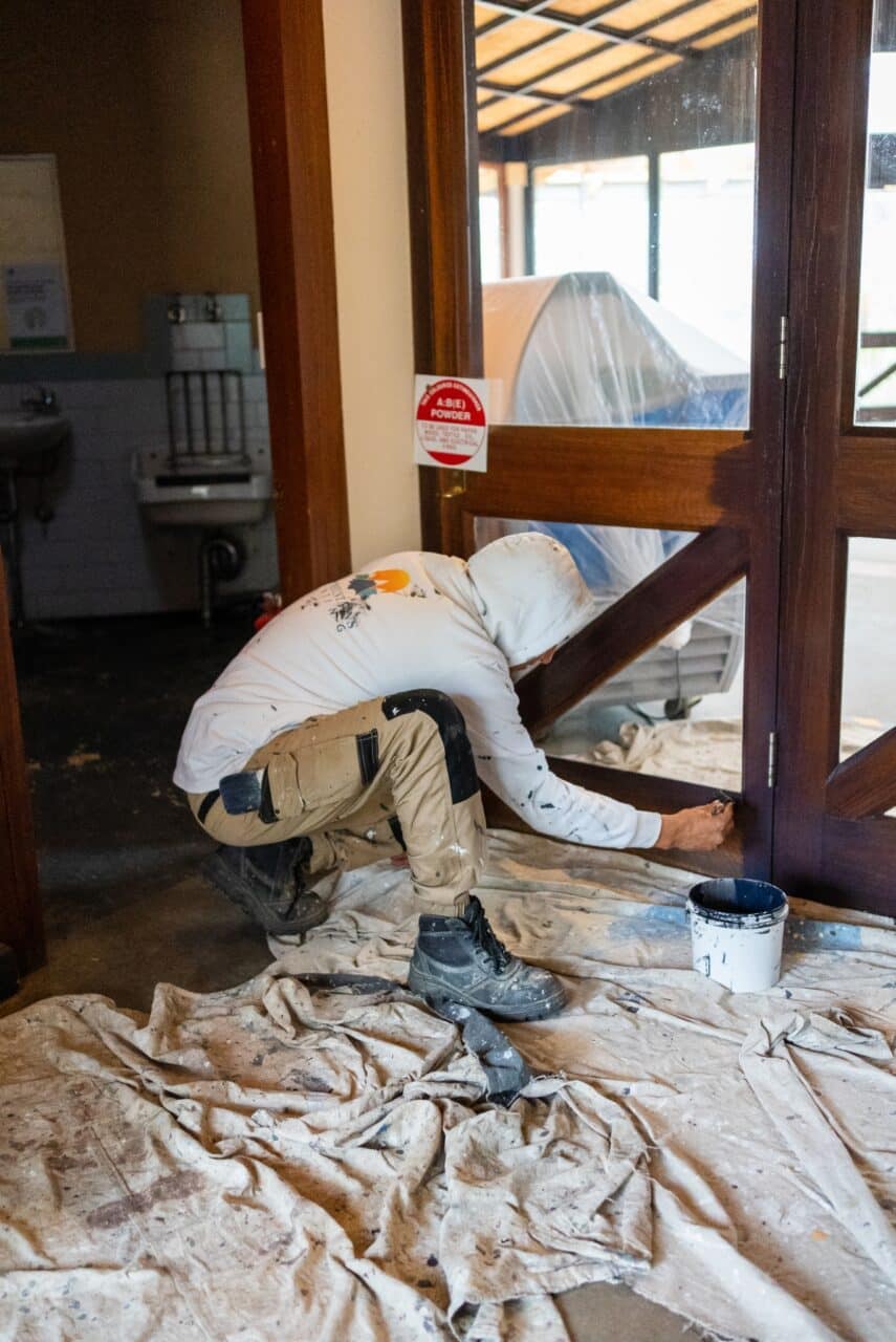 A person in work clothes crouches and paints the lower part of a wooden hut door. Paint supplies are on a drop cloth covering the floor, ensuring careful conservation of the area.