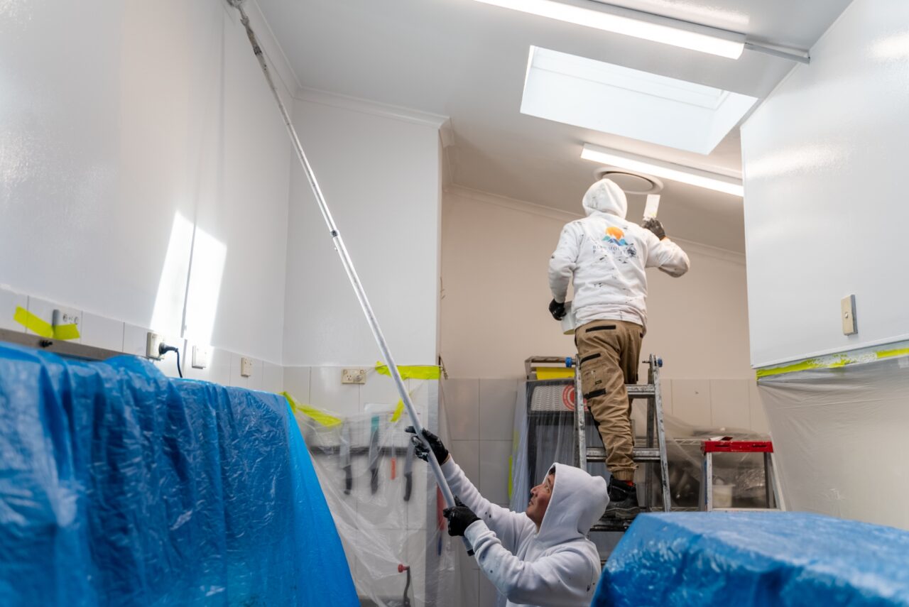 Two painters wearing white hoodies work on painting a high ceiling in a room. One is on a ladder, and the other uses a paint roller on an extended pole. With the furniture covered by protective blue sheets, their efforts contribute to the conservation of this cozy hut-like space.