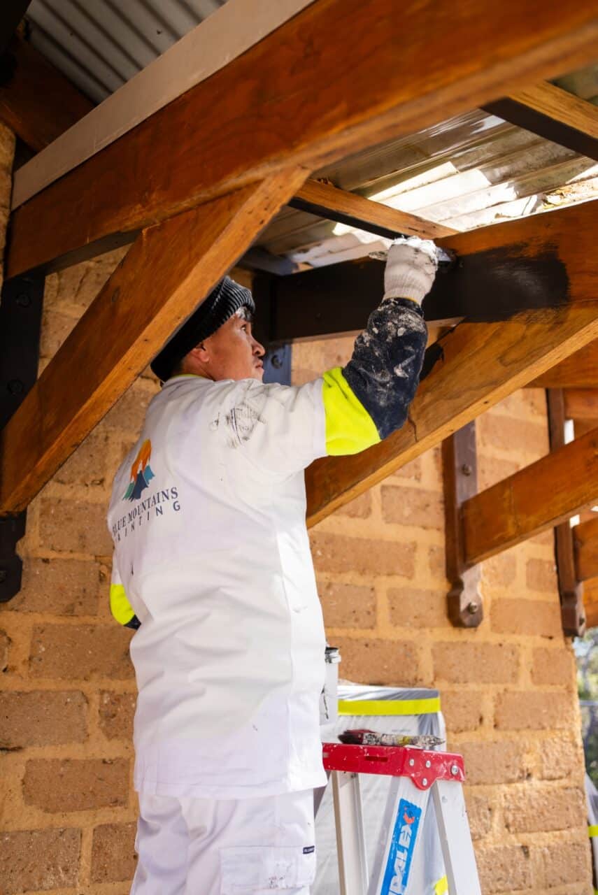 A worker in protective clothing stands on a ladder, painting the wooden beams of the conservation hut's roof structure.